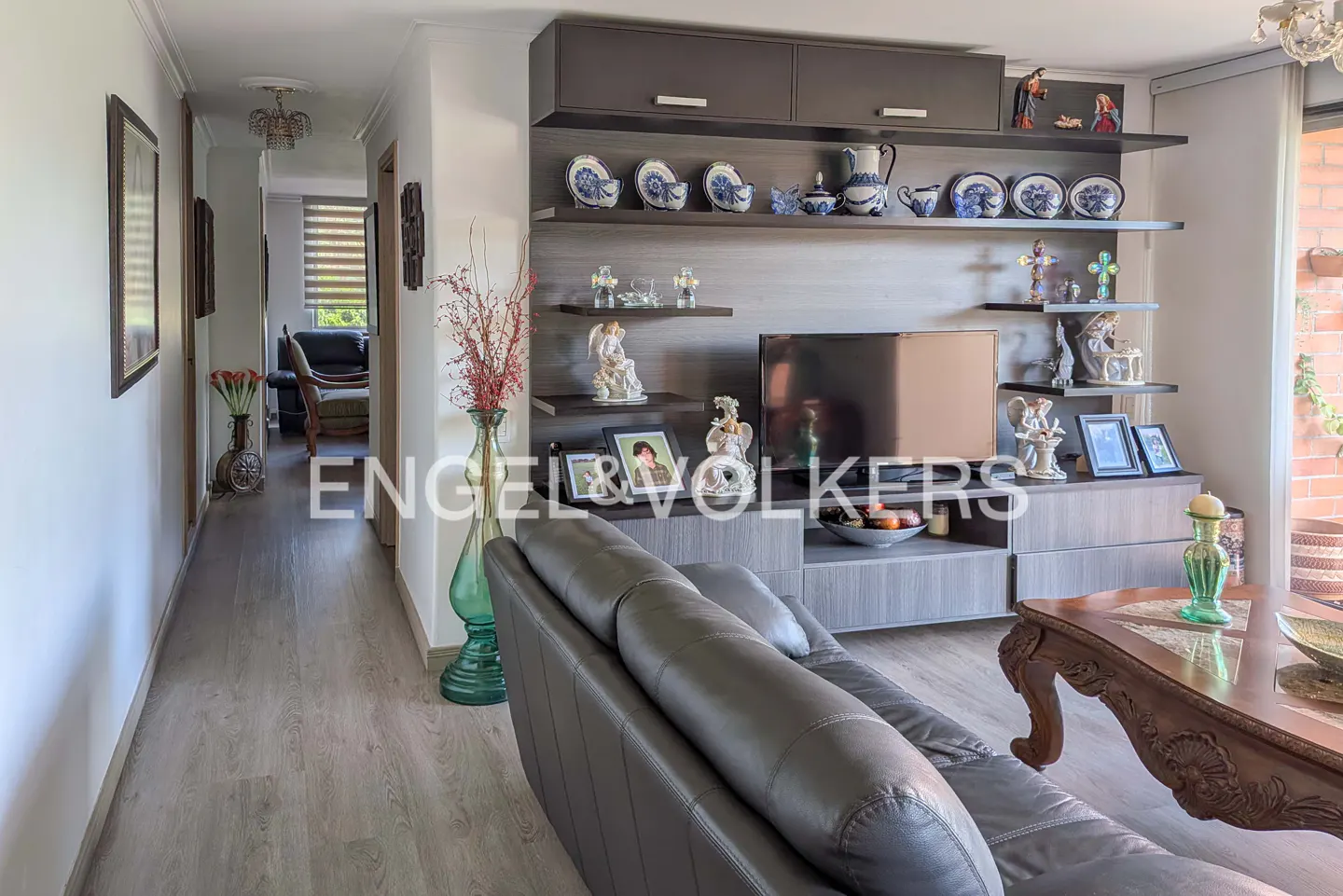 Living room with gray sofa, wood floors, and a wall unit with shelves displaying decorative items and a TV. A hallway leads to other rooms.