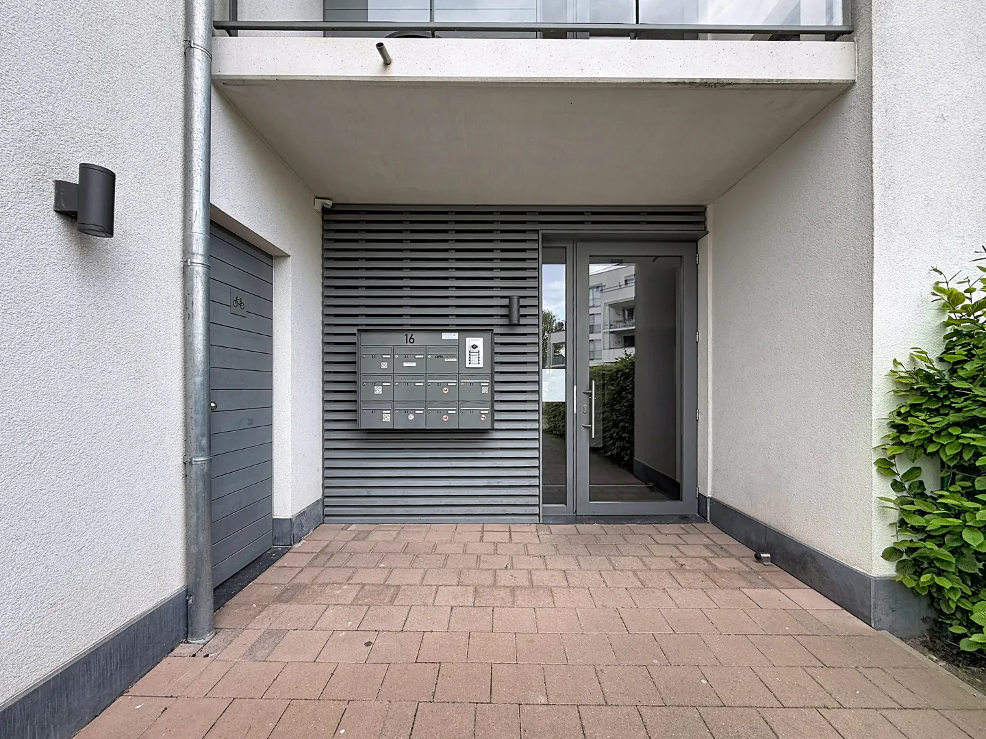 Apartment building entrance with gray door, mailboxes, and brick walkway. White walls and green bushes add contrast.