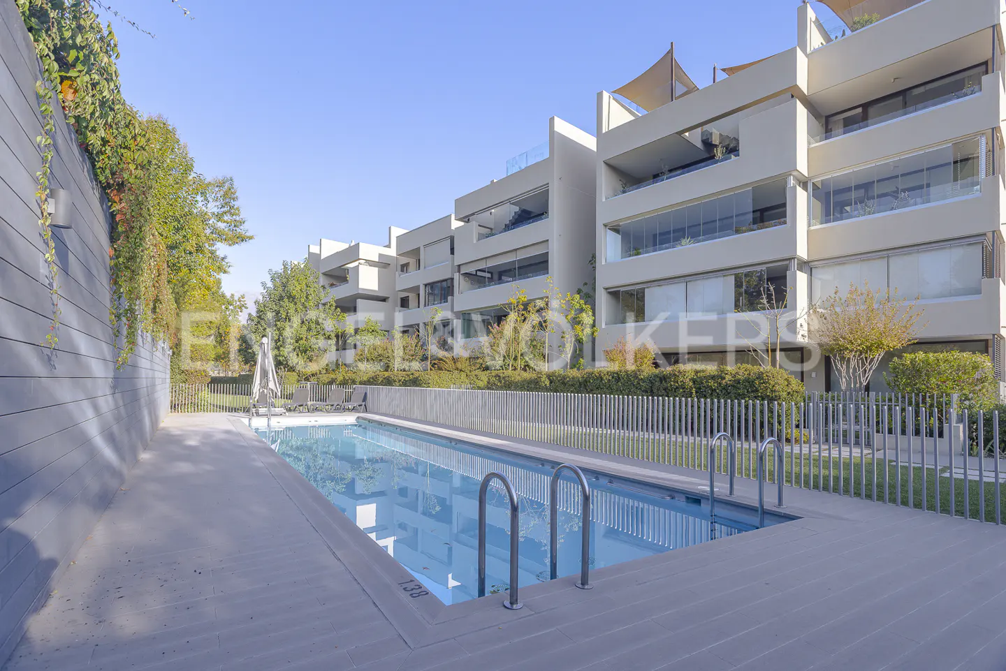 A long, rectangular swimming pool with metal ladders, next to modern apartments under a blue sky.