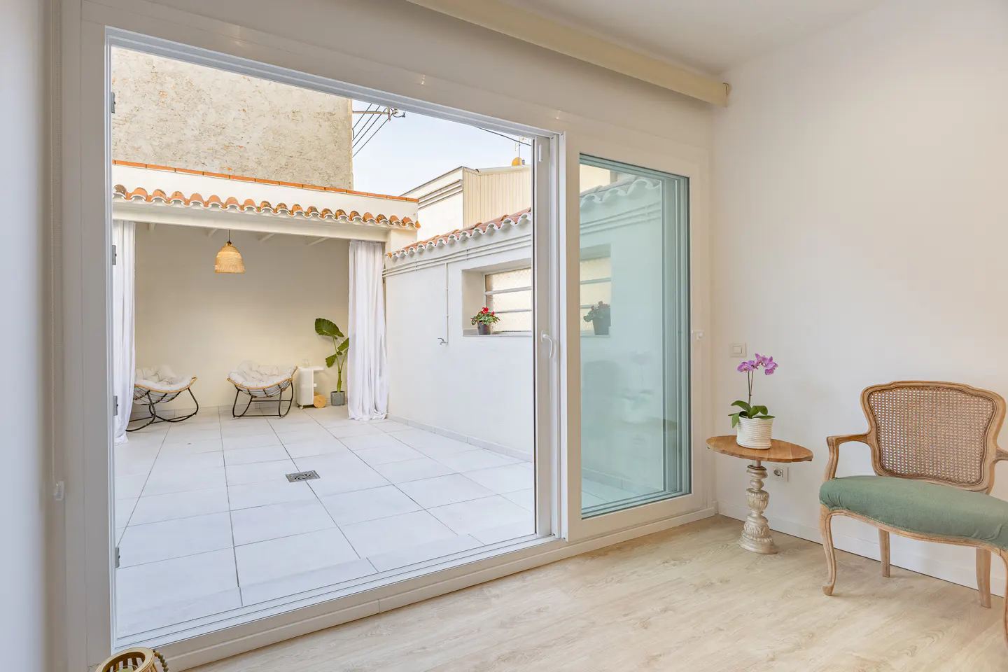 Bright room with a sliding glass door to a white-walled patio with chairs, a plant, and a hanging light. A green chair and orchid are inside.