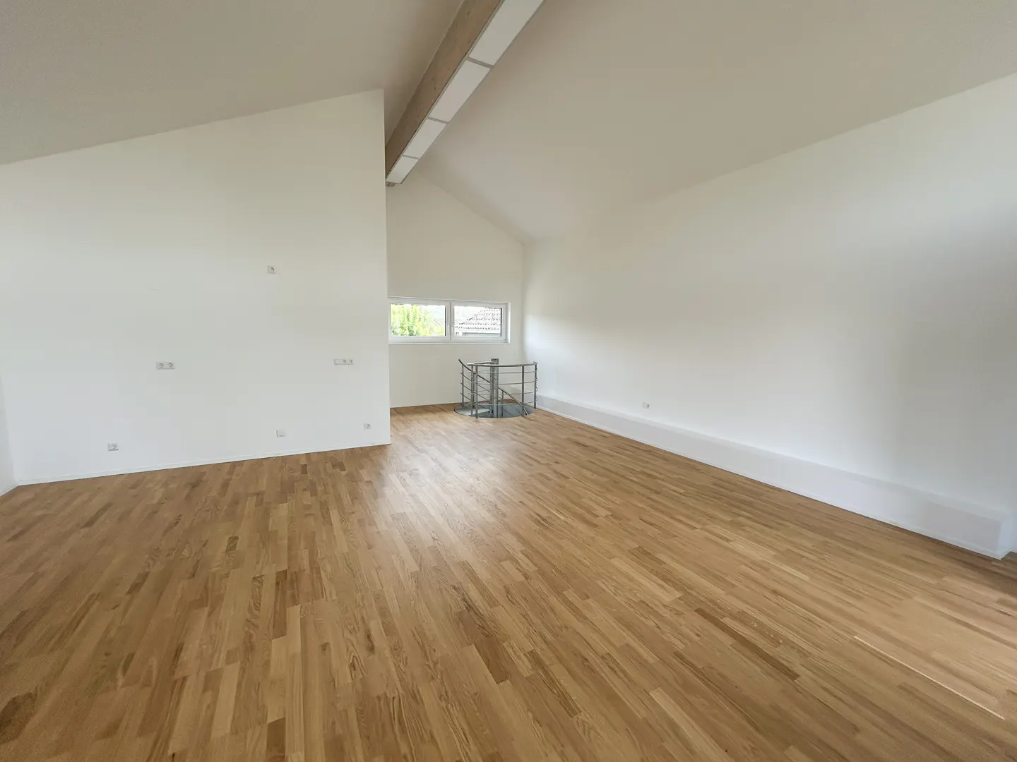 Spacious, empty room with white walls, vaulted ceilings, light wood flooring, and a small metal spiral staircase.