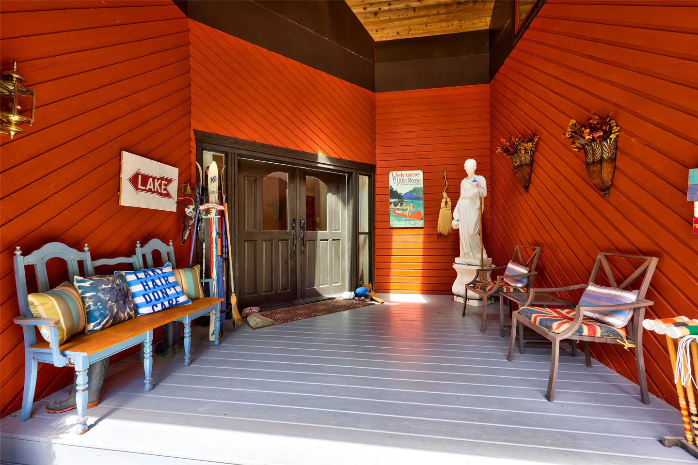 Inviting porch with orange wood walls, gray floor, and rustic decor. Bench, chairs, skis, and a "Lake" sign add charm. Double doors in the center.
