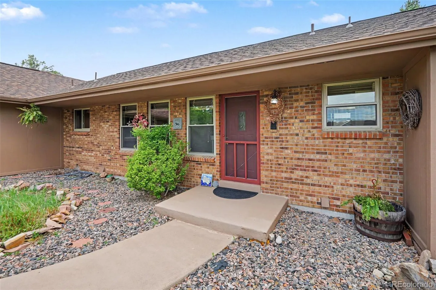 A single-story brick house with a red door, a concrete walkway, and rock landscaping.