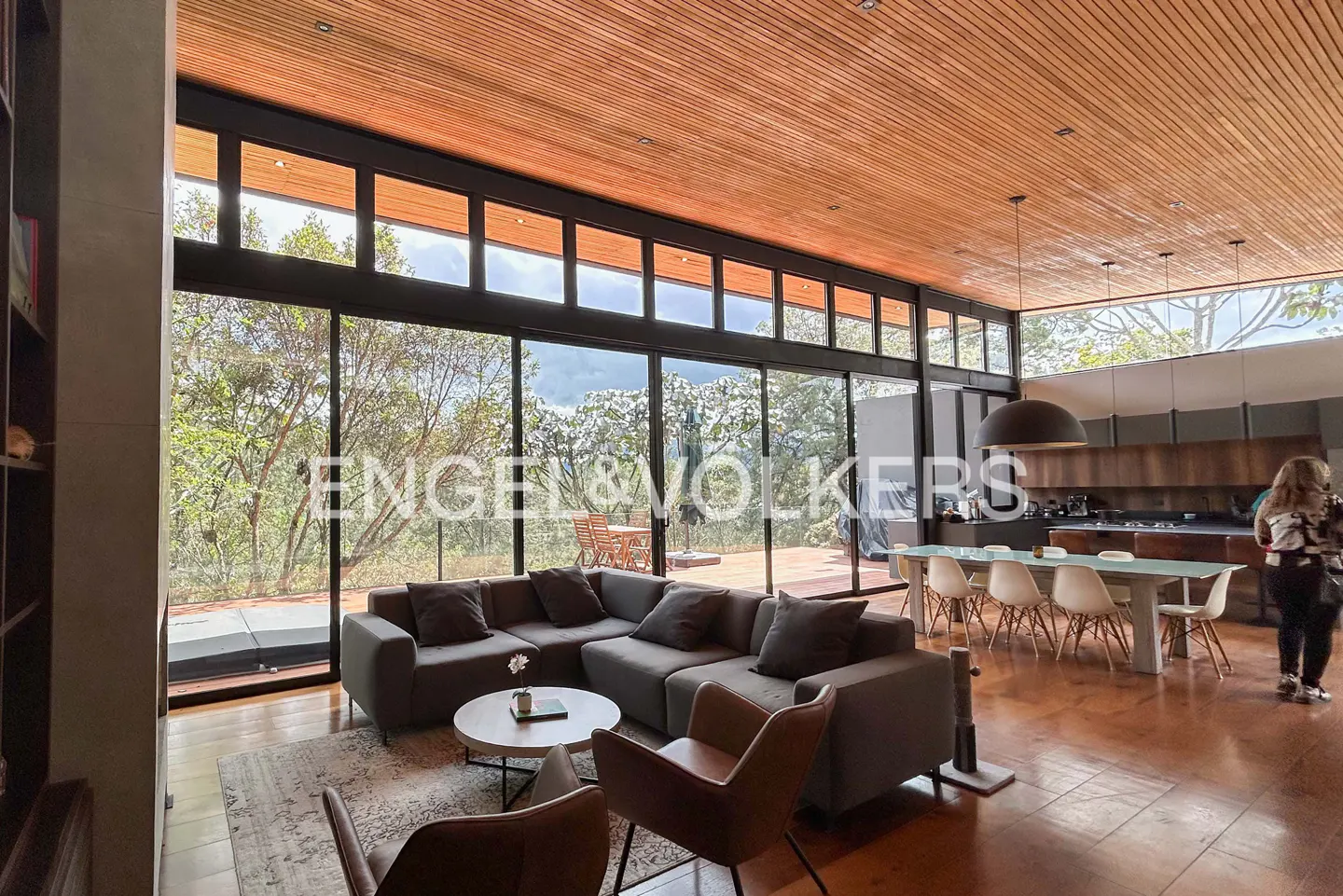 Open-concept living space with wood ceiling, floor-to-ceiling windows, gray sectional sofa, and dining table with white chairs.