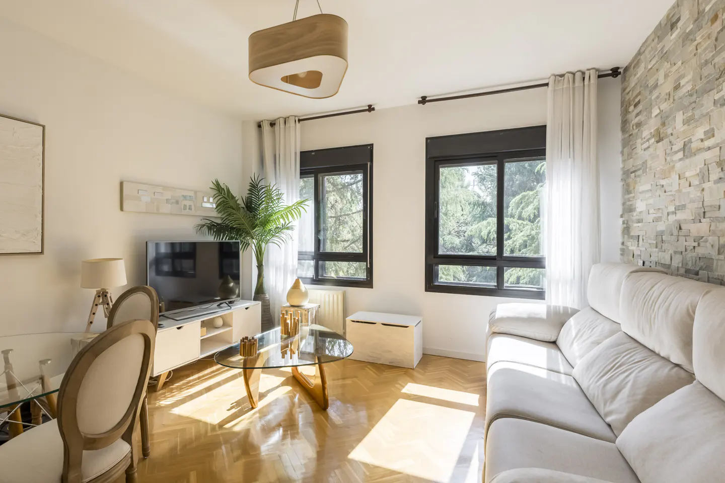 Bright living room with wood floors, white sofa, glass table, TV, and two windows with black frames. A stone accent wall adds texture.