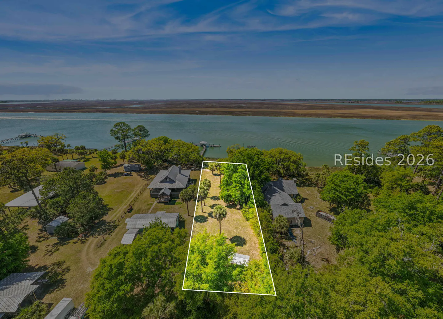 Aerial view of a waterfront property outlined in white, with trees, houses, and a blue sky.