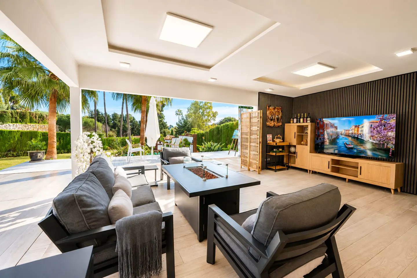 Bright, modern patio with gray furniture, a black fire pit table, and a view of a green lawn with palm trees. Inside, a TV displays a Venice canal scene.