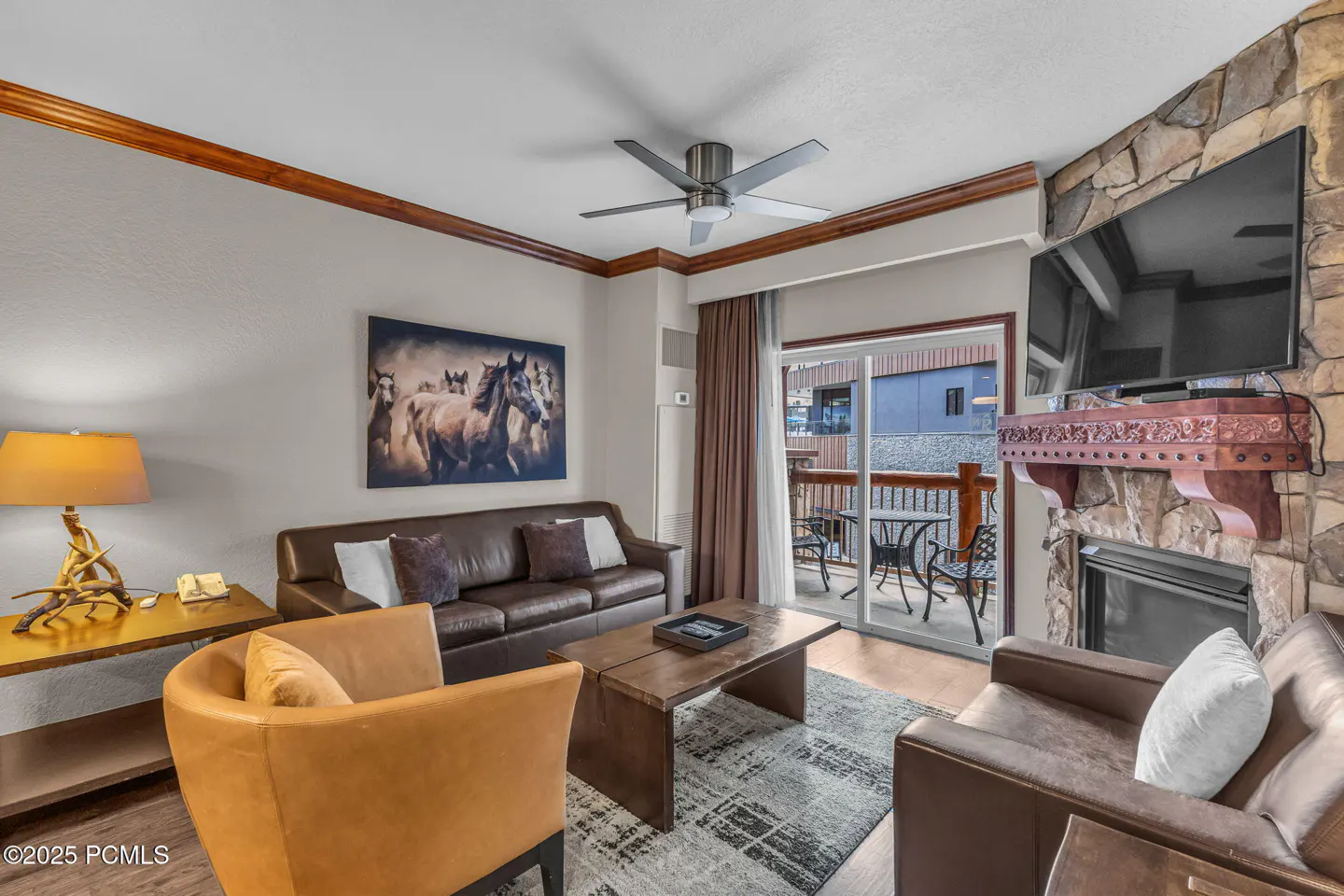 Living room with brown leather sofa, tan chair, stone fireplace, and balcony with outdoor seating.