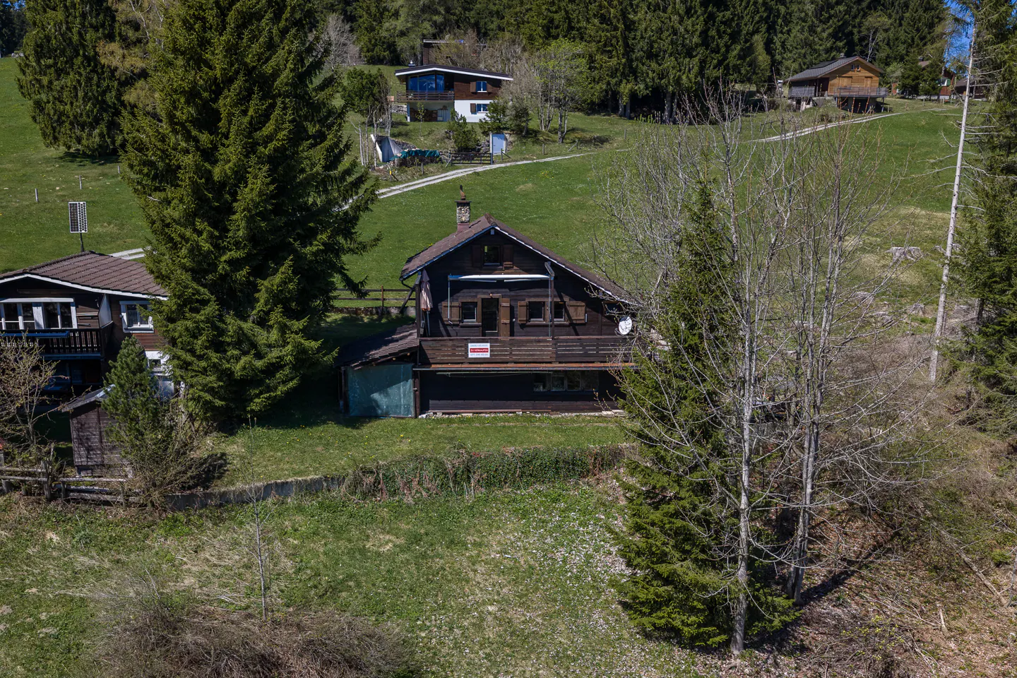 A brown chalet-style house with a balcony, nestled on a green hillside with trees.