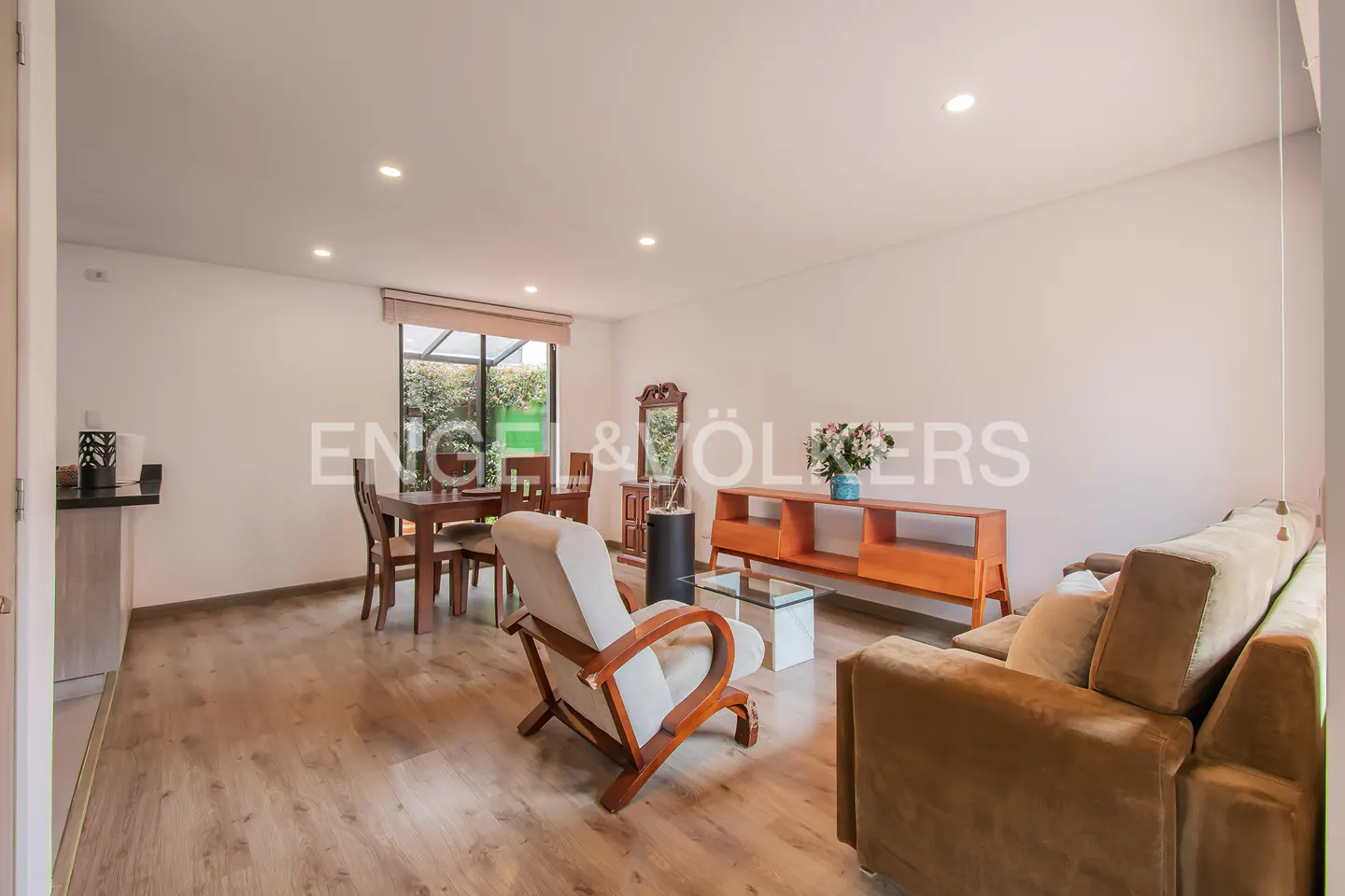 Bright living room with wood floors, white walls, and recessed lighting. A brown sofa, chair, table, and dining set furnish the space.