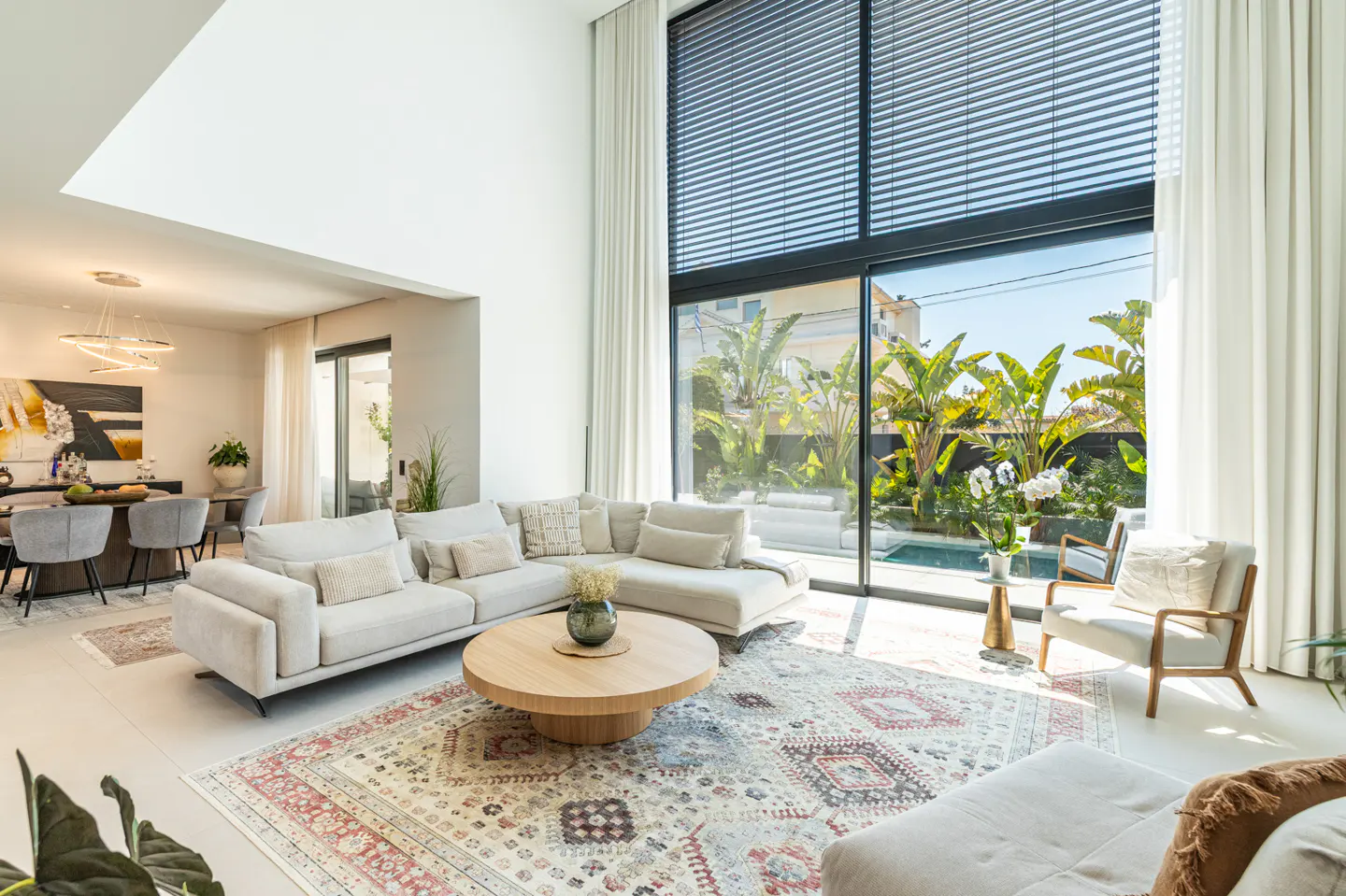 Bright, modern living room with a beige sectional sofa, round wood table, and patterned rug. Large windows overlook a pool and lush greenery.