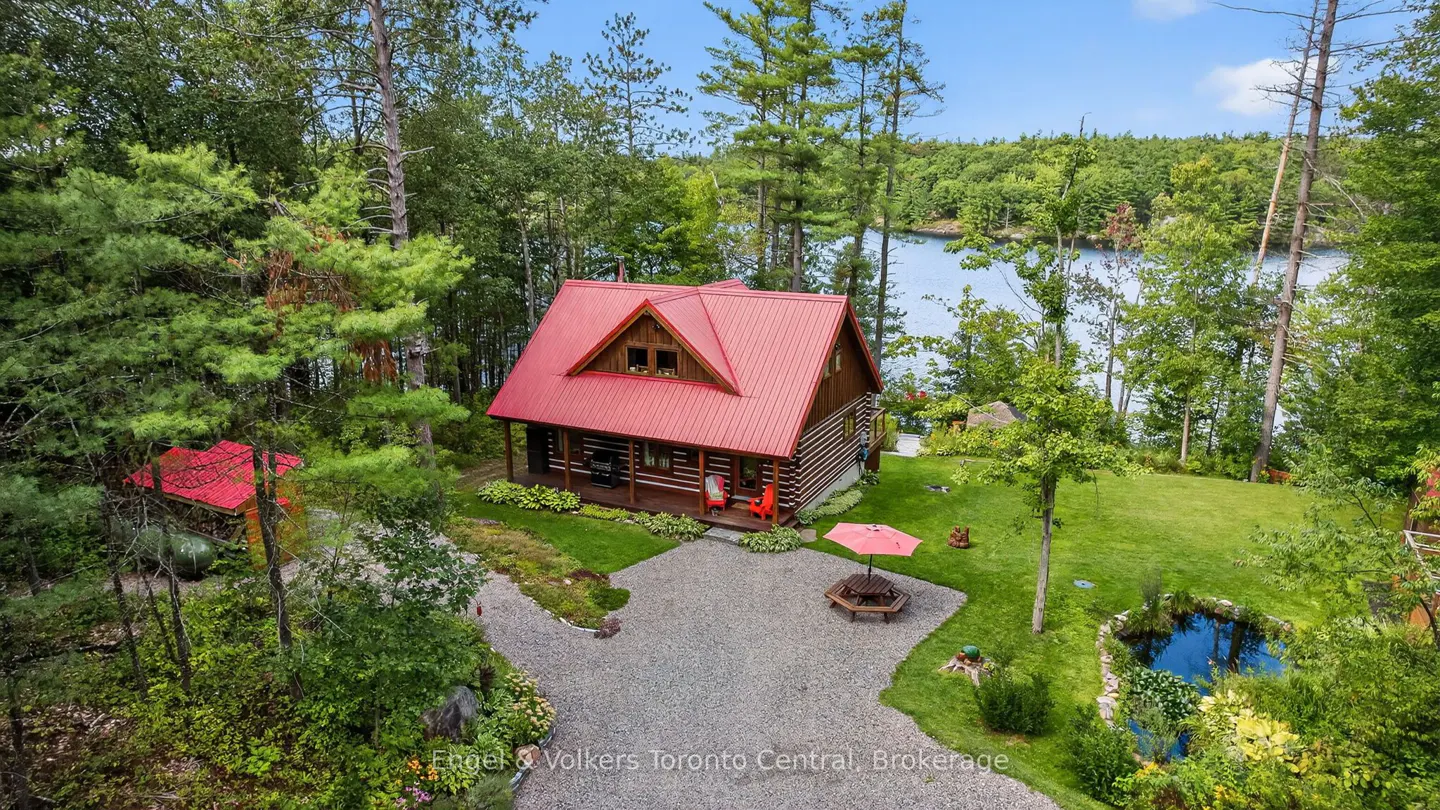 Aerial view of a log cabin with a red roof, surrounded by trees and a lake in the background. A gravel driveway leads to the cabin.