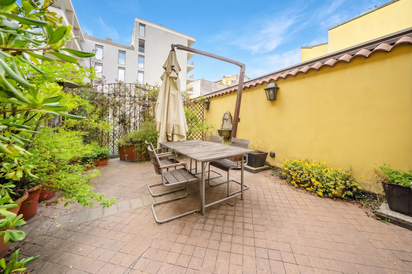 Outdoor patio with a wooden table and chairs, surrounded by greenery and a yellow wall with a fountain. Blue sky above.