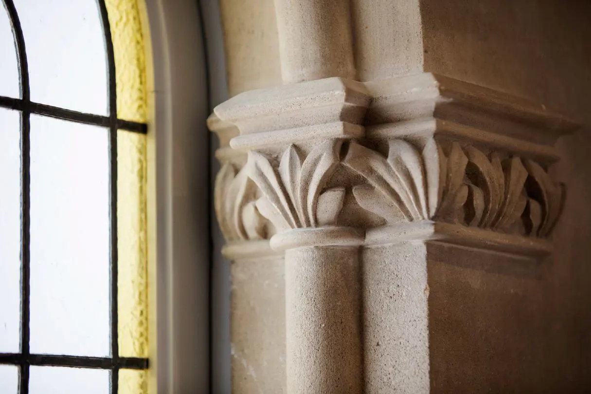 Close-up of a beige stone column with carved leaf details, next to a window with black grids and a yellow frame.