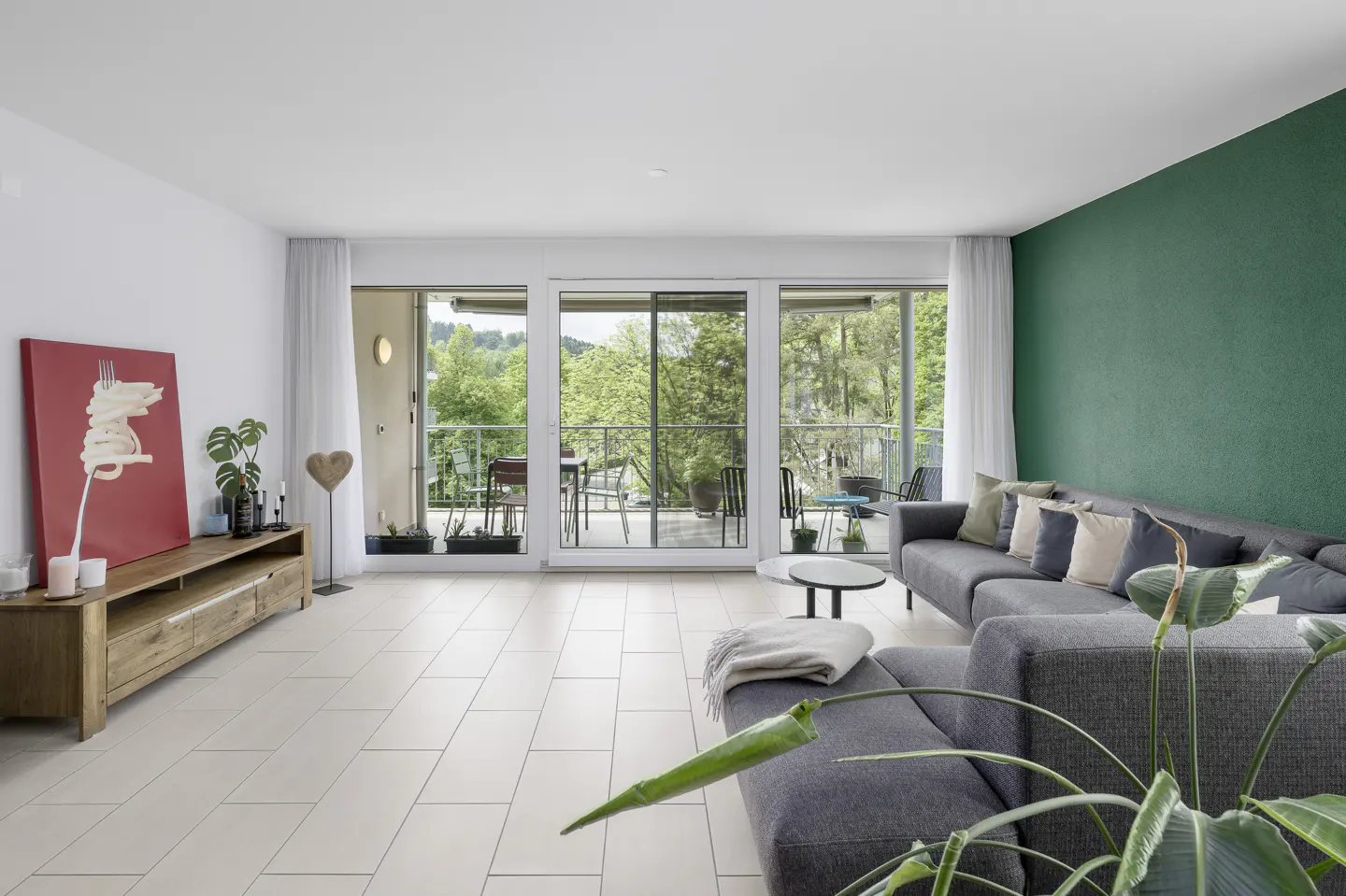 Bright living room with a gray sectional sofa, green accent wall, and balcony with outdoor seating. Artwork and plants add pops of color.