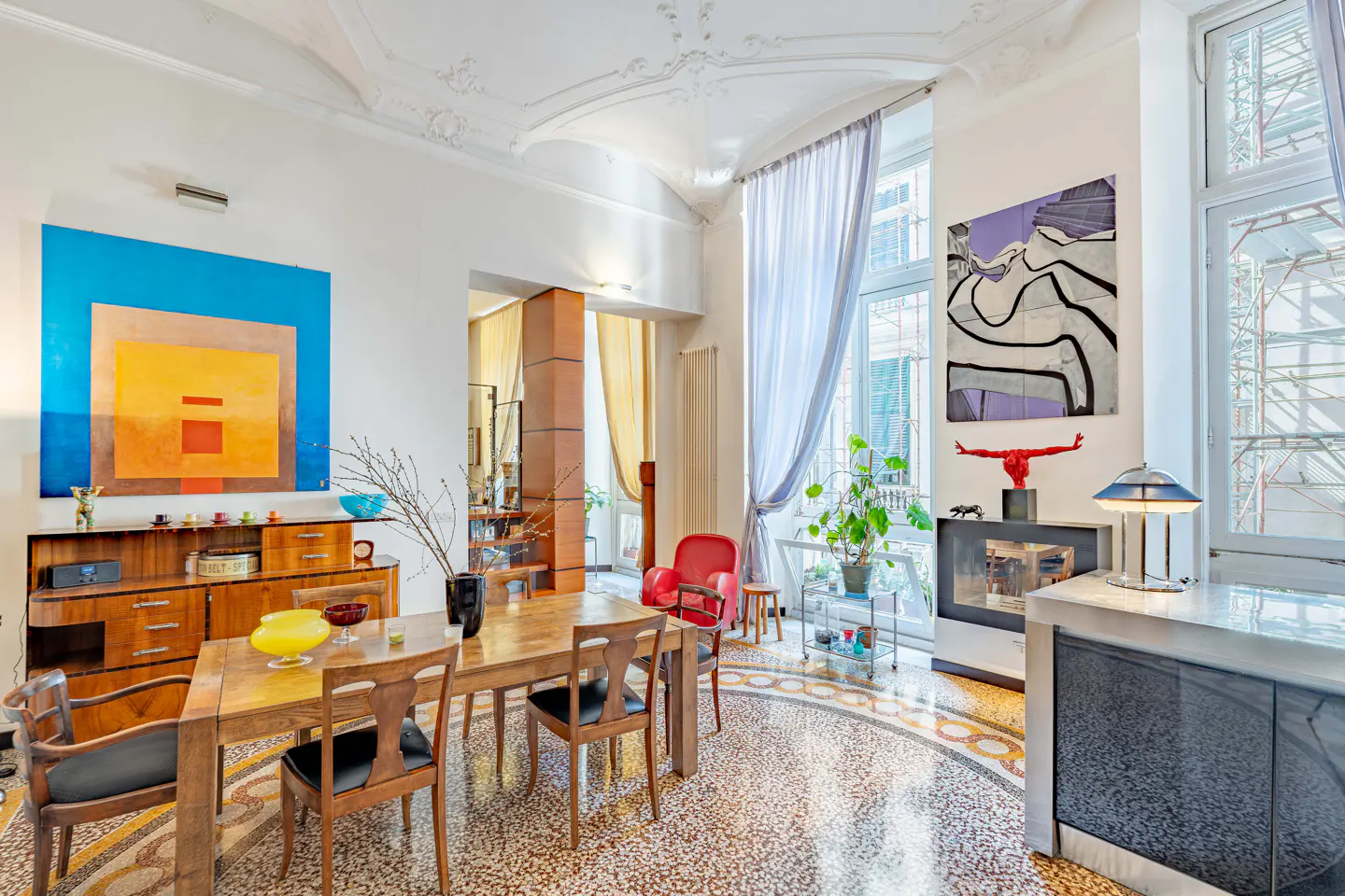 Bright, eclectic dining room with wood table, chairs, and cabinet. Large colorful paintings adorn the white walls. Mosaic tile floor. Curtained windows.