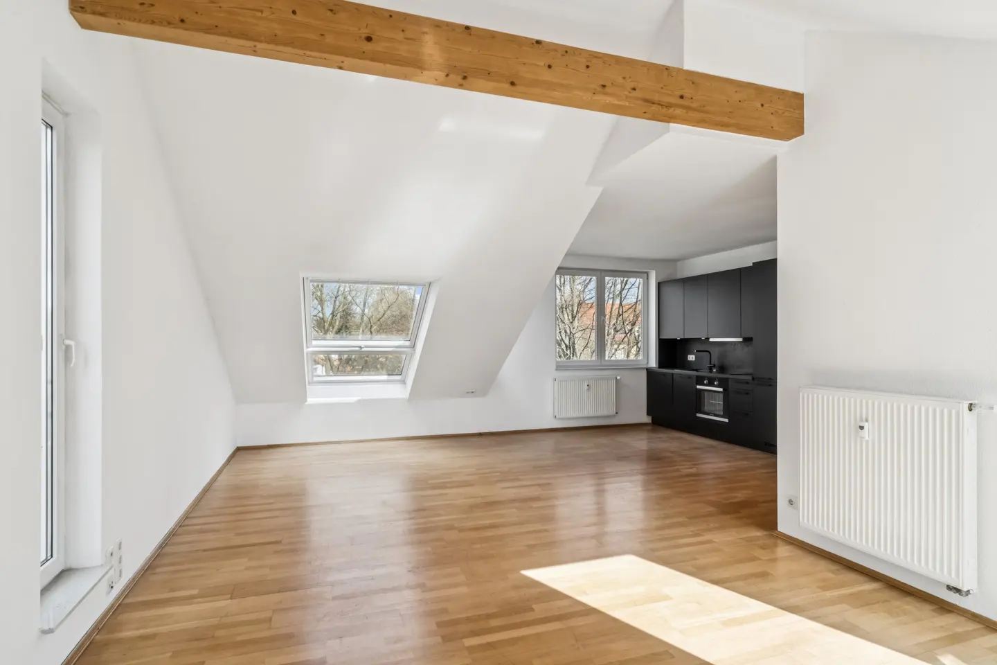 Bright, empty attic apartment with wood floors, white walls, and a black kitchen. A wooden beam crosses the ceiling. Sunlight streams through windows.