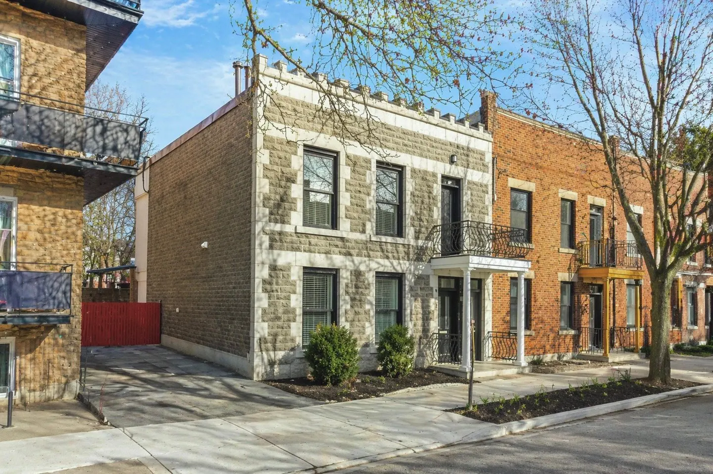 Two-story stone house with black windows and a small balcony, next to a brick house on a sunny street.