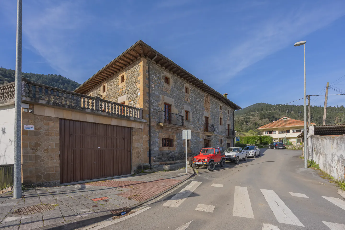 A stone house with a brown garage door sits on a street with parked cars and a crosswalk.