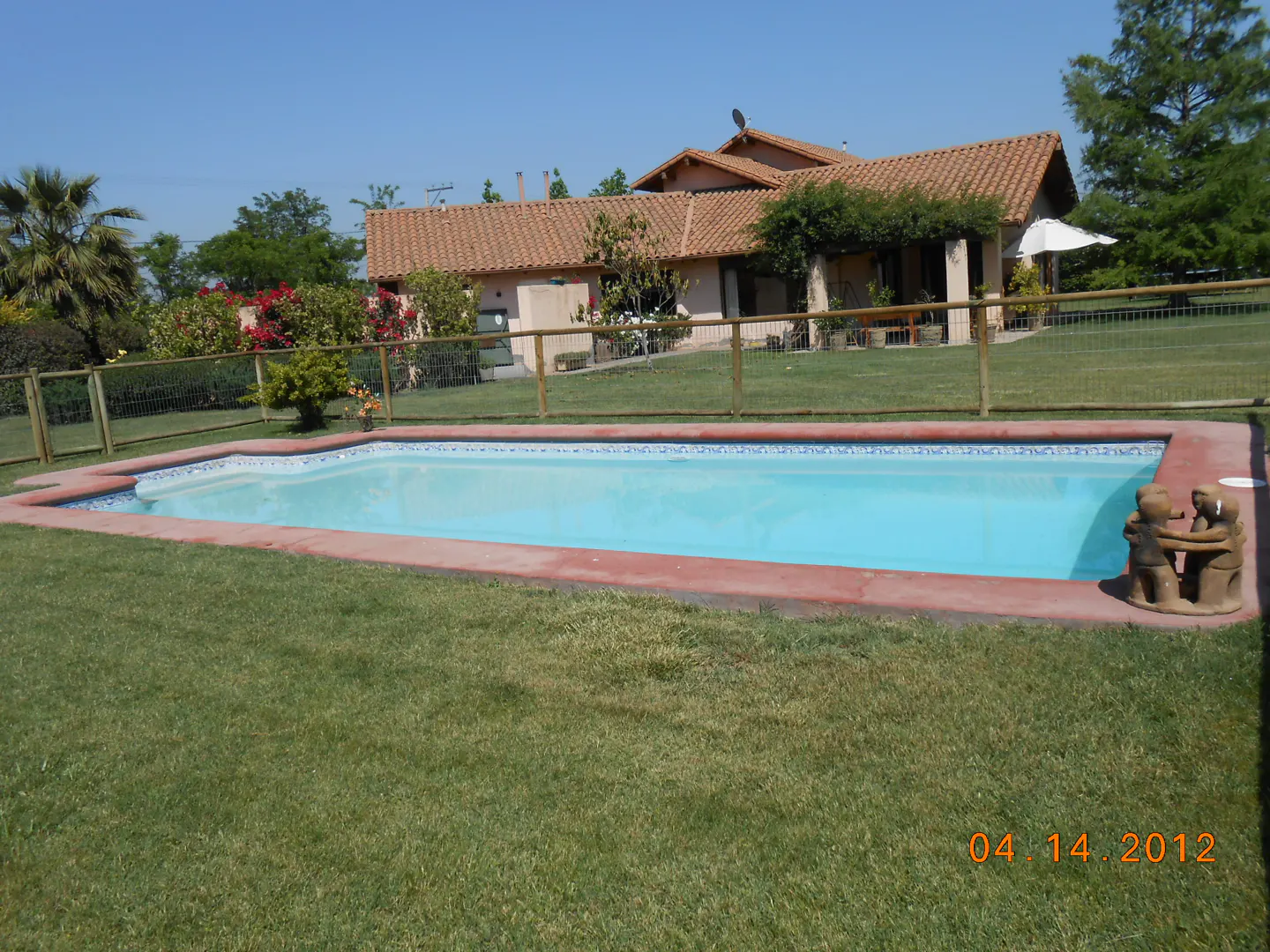 Outdoor pool with red trim and blue tile, surrounded by green grass and a wooden fence. A house with a red tile roof is in the background.
