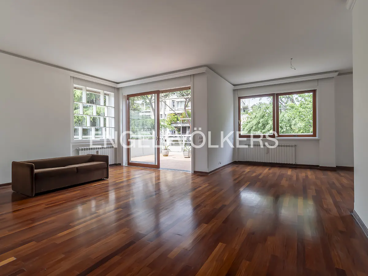 Bright, empty living room with hardwood floors, white walls, and a brown sofa. Sliding glass doors lead to a balcony. Windows offer views of greenery.