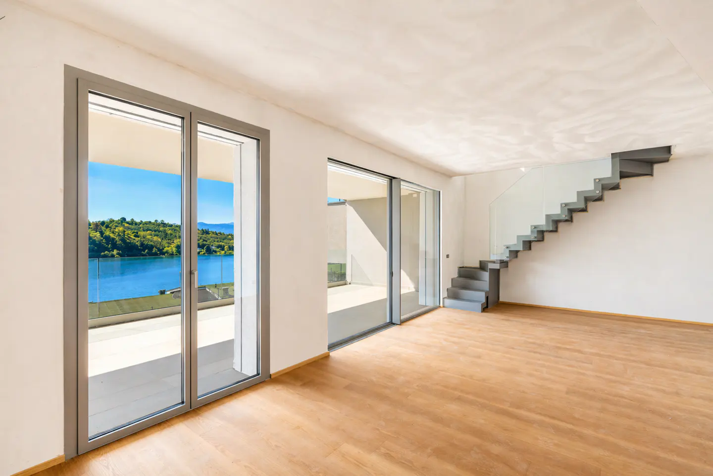 Bright, empty room with wood floors, white walls, and gray stairs. Sliding glass doors open to a lake view.