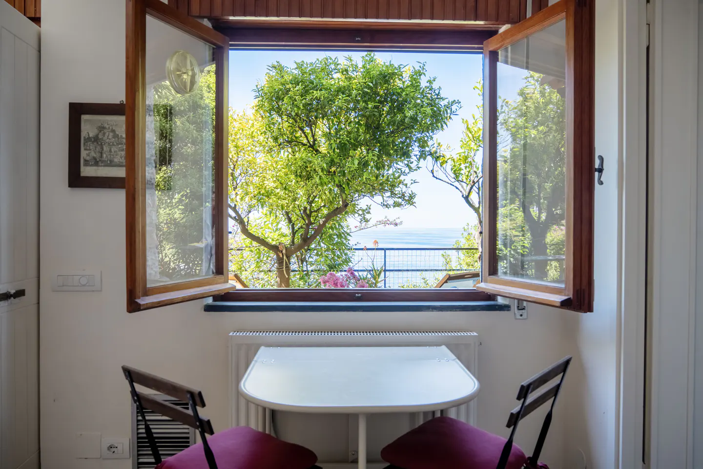Interior view of a white room with an open window framing a lush green tree and a distant blue sea. A small white table and two chairs sit in front of the window.