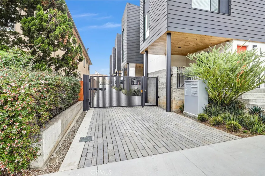 Modern townhouses with gray siding and black metal gates, viewed from a gray brick driveway. Green bushes and trees surround the property.