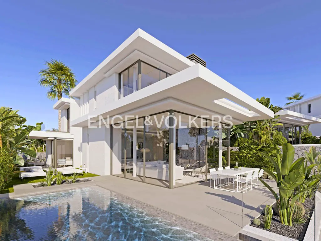 Modern white house with glass walls, a pool, and patio furniture under a blue sky surrounded by lush greenery.