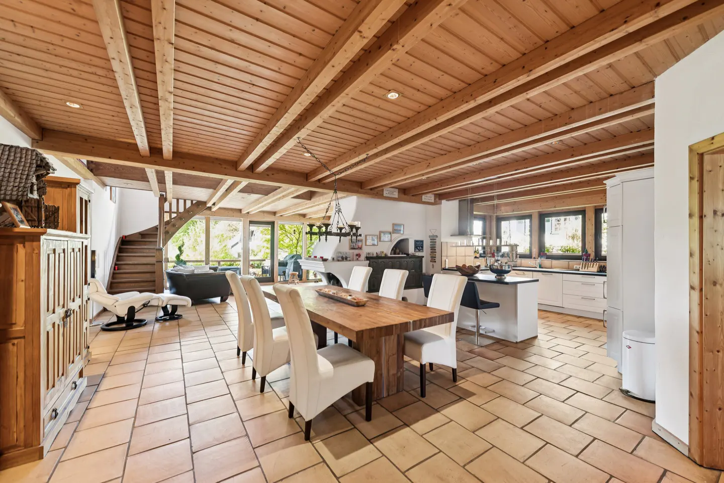 Open-plan dining area with a wooden table, white chairs, tiled floor, and exposed wooden ceiling beams.