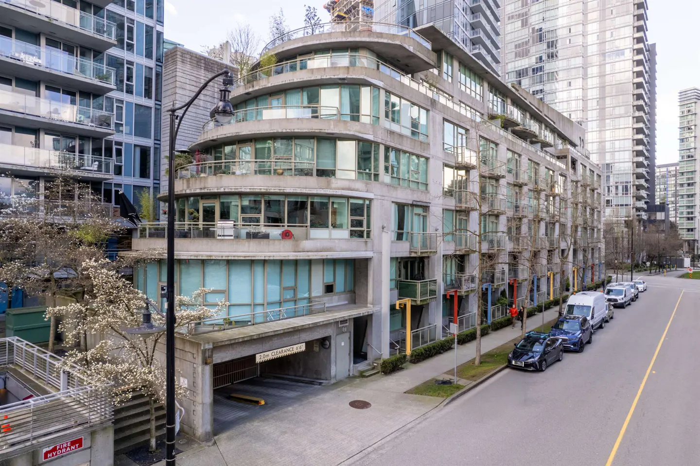 Exterior view of a modern, multi-story concrete apartment building with curved balconies and a street with parked cars.