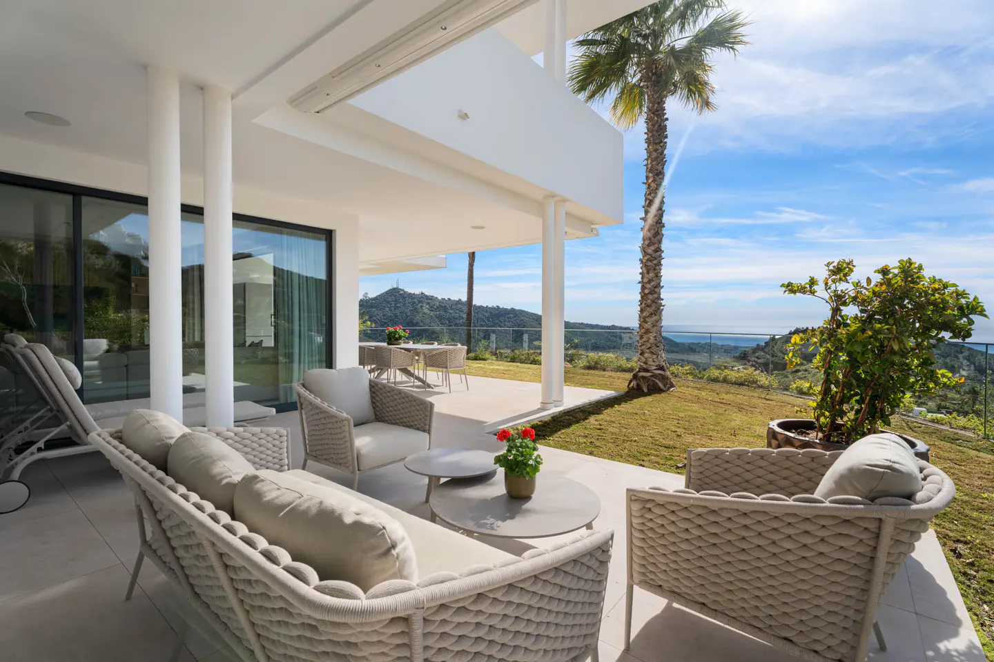 Outdoor patio with wicker furniture, a palm tree, and a view of the mountains and ocean under a blue sky.