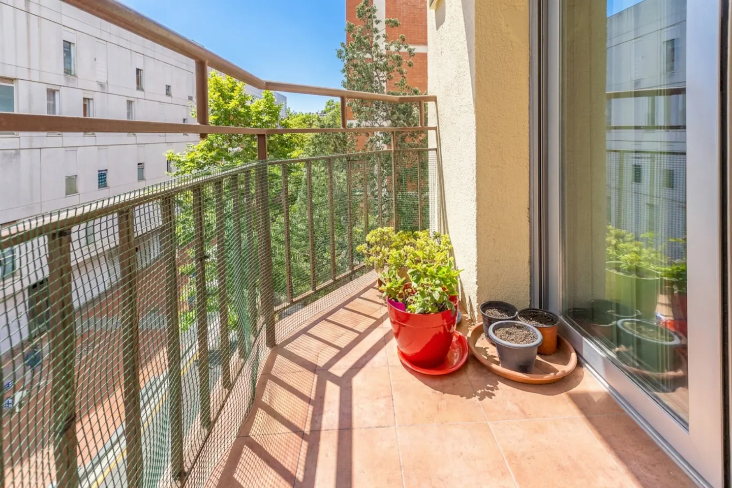 Small balcony with tiled floor, metal railing, potted plants, and a view of a neighboring building on a sunny day.