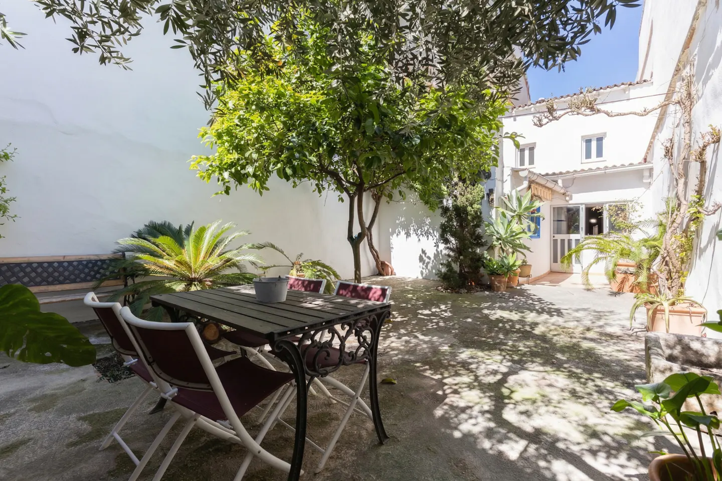 Outdoor patio with a wooden table and chairs, surrounded by lush greenery and a white building in the background.