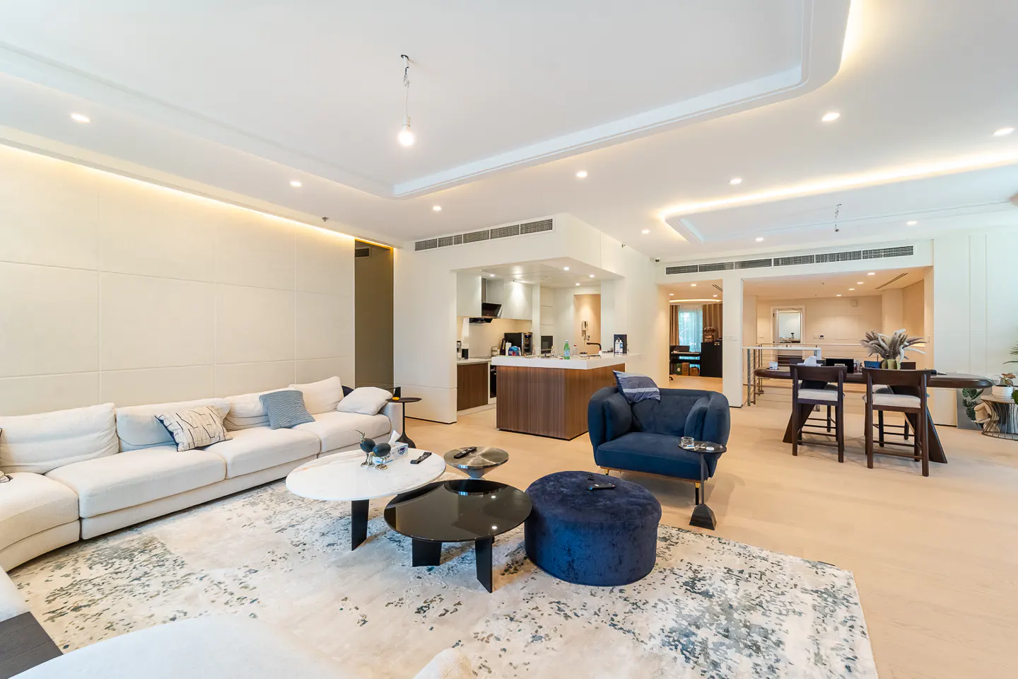 Bright, open-concept living space with a white sofa, blue chair, and round tables on a patterned rug. Kitchen and dining area visible in the background.