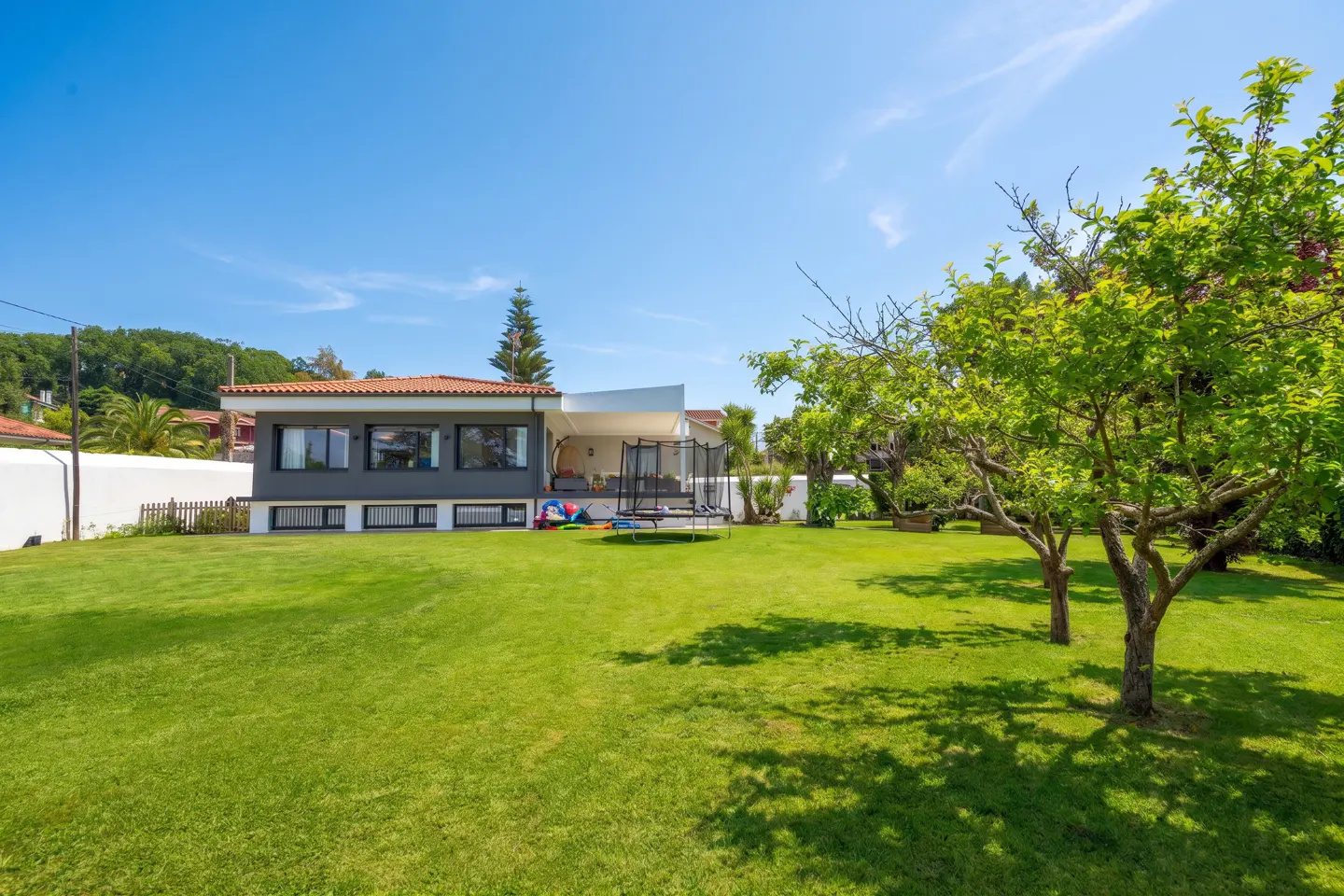 A modern gray house with a red tile roof overlooks a large green lawn with trees and a trampoline under a clear blue sky.