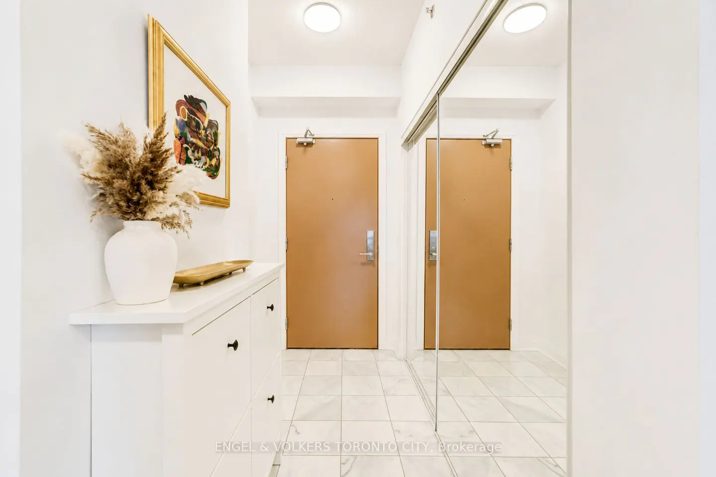 Hallway with white walls, marble floors, and a white cabinet with a vase of pampas grass. A framed painting hangs above the cabinet. Two brown doors and a mirrored closet are visible.