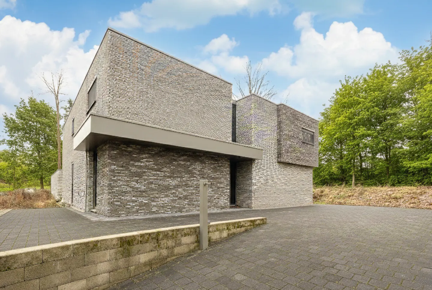 Modern gray brick house with a flat roof and a paved driveway, surrounded by green trees under a cloudy blue sky.