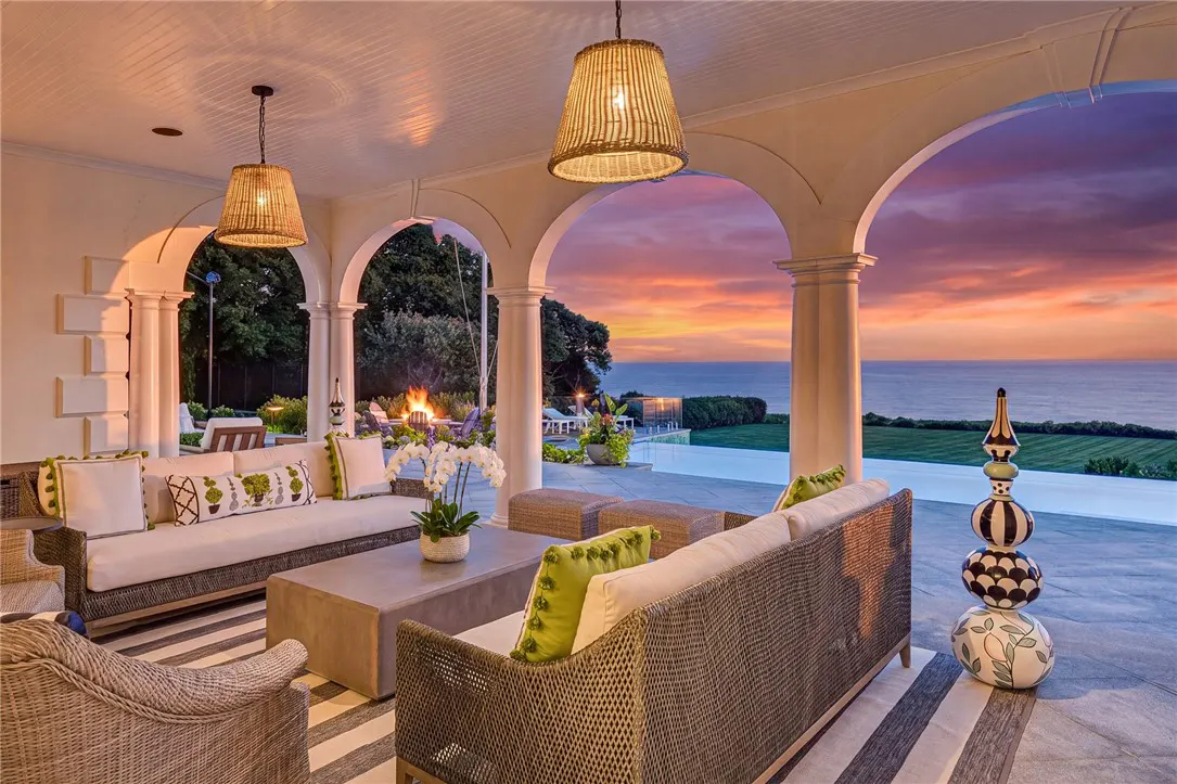 Covered patio with wicker furniture, white cushions, and green accents. Arches frame an ocean view with a vibrant sunset.
