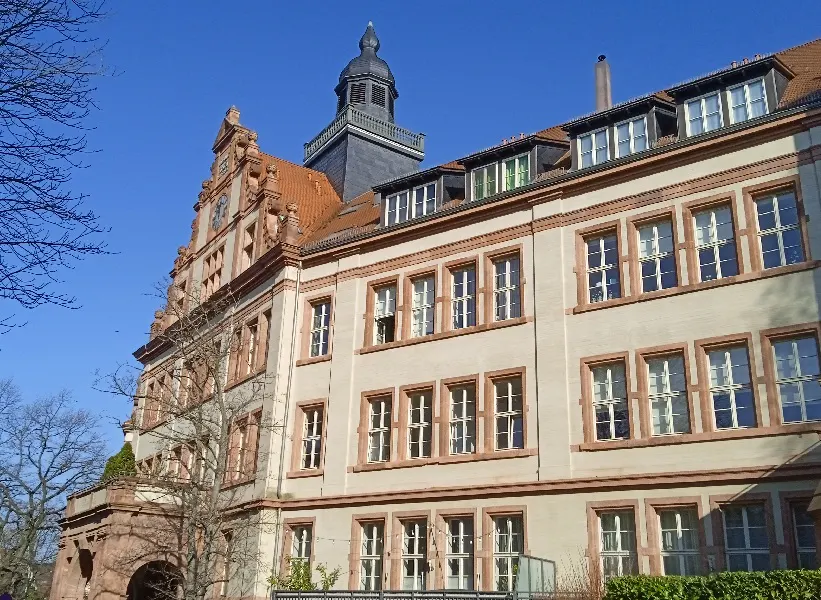 Exterior view of a large, light-beige building with red trim and a dark-gray tower under a clear blue sky.
