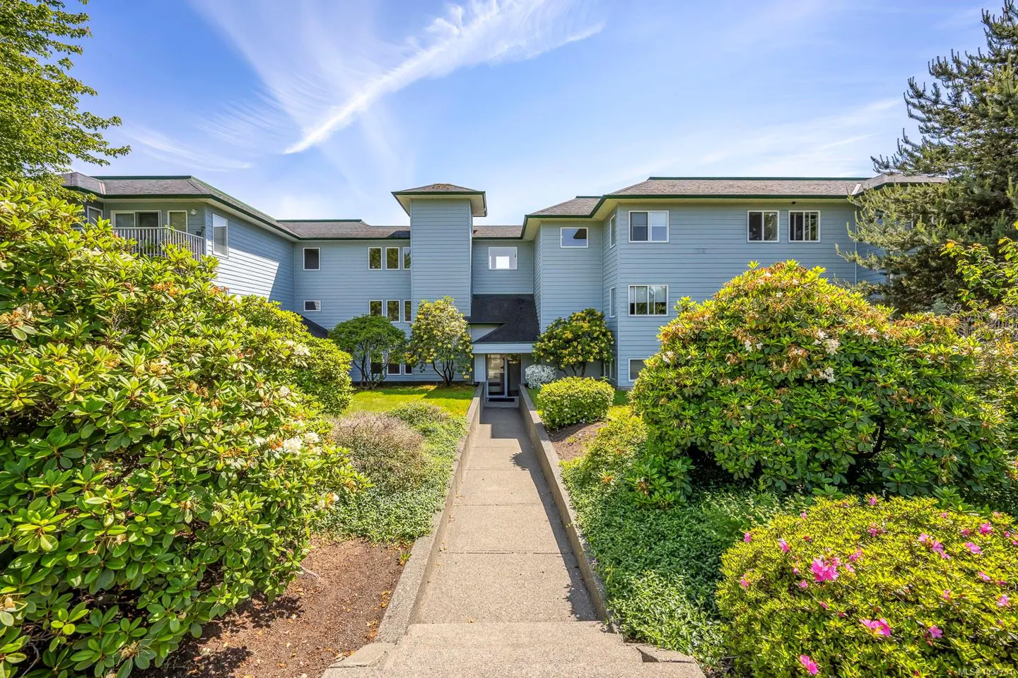 Exterior view of a light blue apartment building with a concrete walkway leading to the entrance, surrounded by green bushes.
