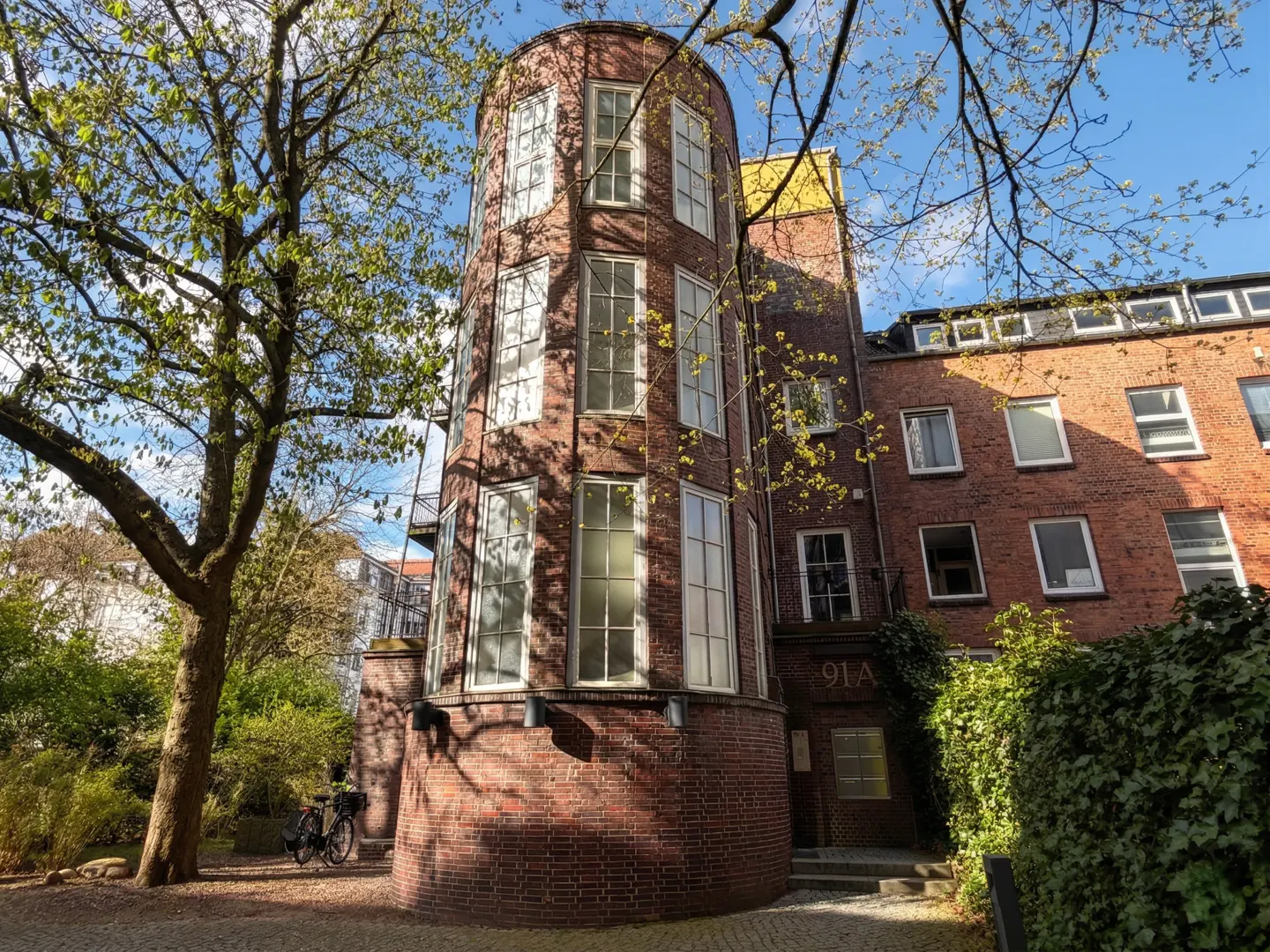 Exterior view of a red brick building with a cylindrical tower and many windows, next to a tree and green bushes.