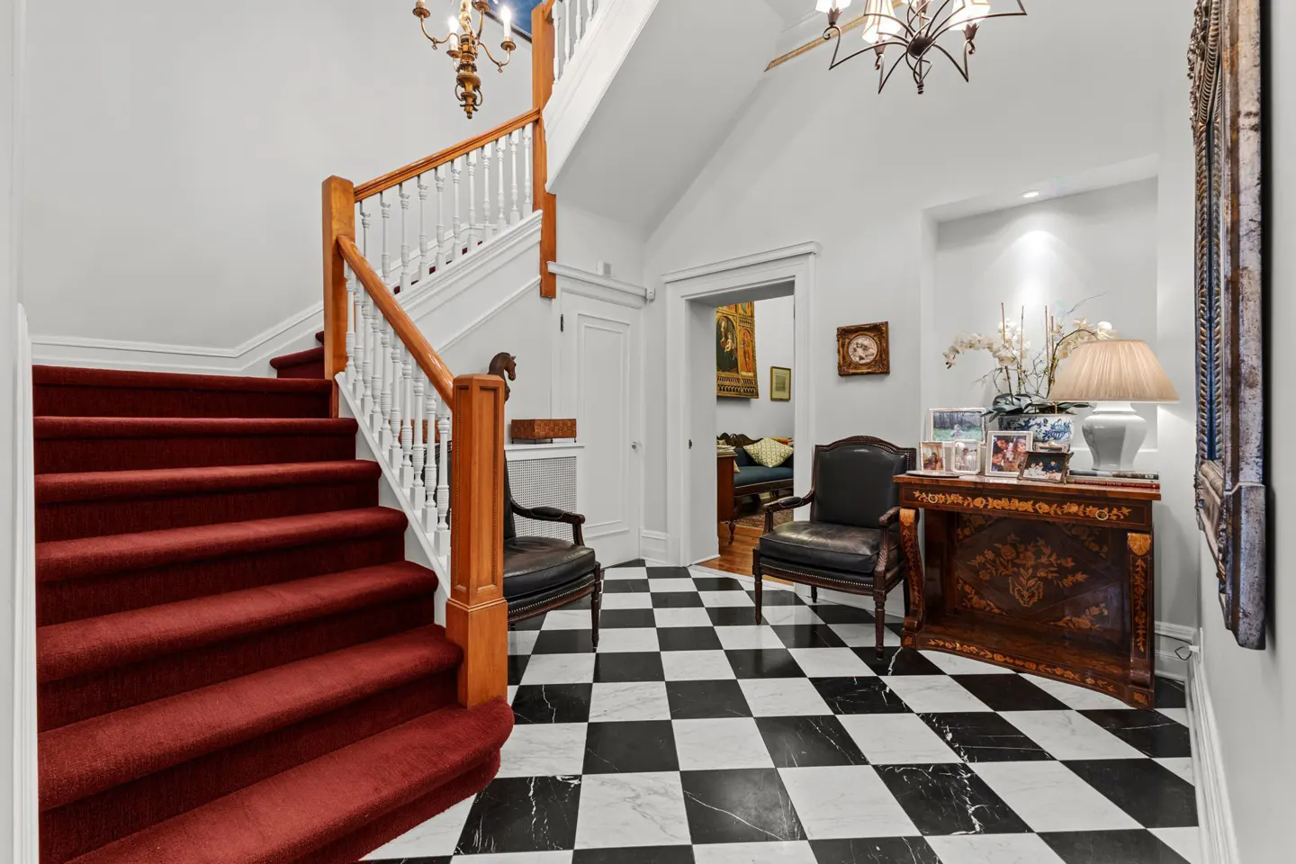 Foyer with red carpeted stairs, black and white checkered floor, two black chairs, and a decorative table with a lamp.