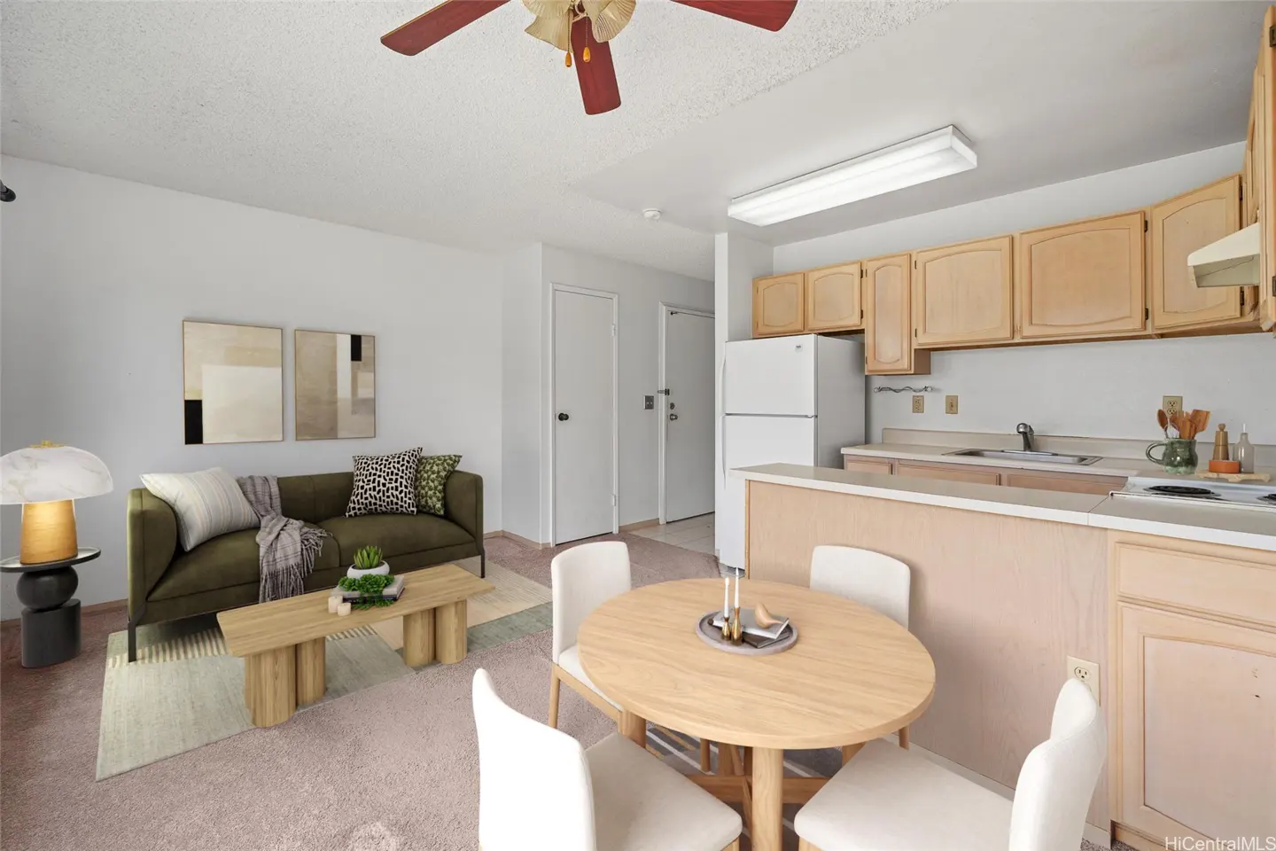 A bright living space with a green sofa, wood table, and a round dining table with white chairs. The kitchen has light wood cabinets and white appliances.