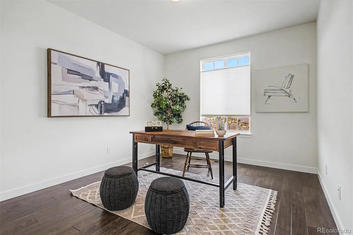 Bright home office with white walls, dark wood floors, and a patterned rug. A wooden desk sits in front of a window, with two gray stools in front.