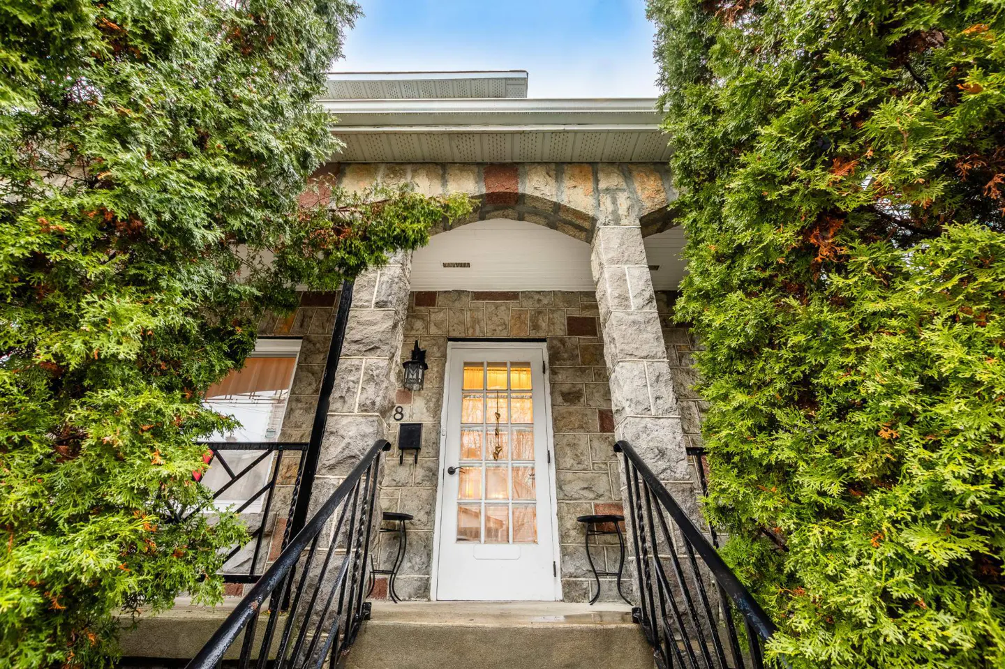 Stone house entrance with white door, black railings, and green trees. The house number "8" is visible next to a black mailbox.