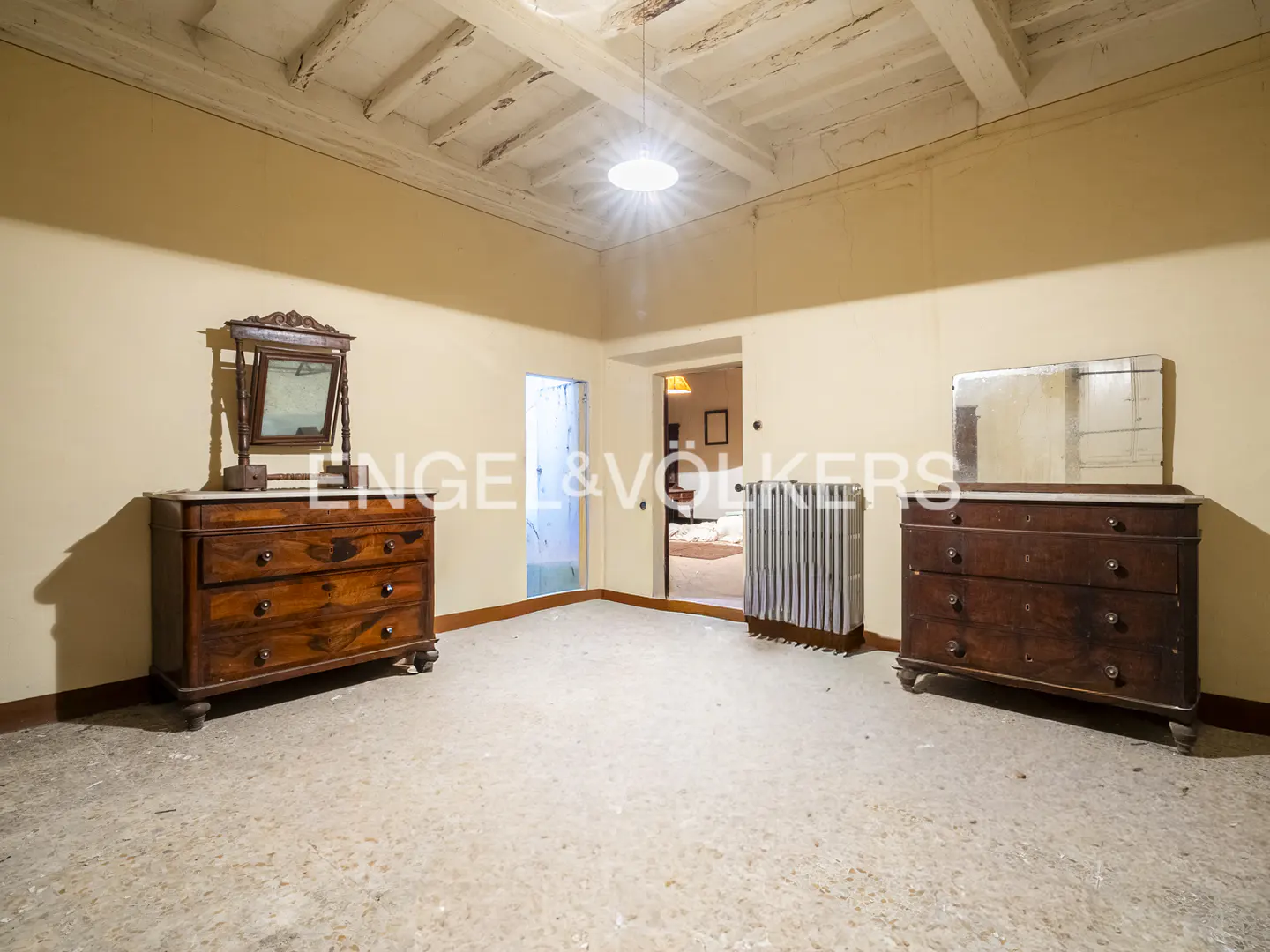 A room with two wooden dressers, mirrors, a radiator, and a white ceiling with exposed beams.