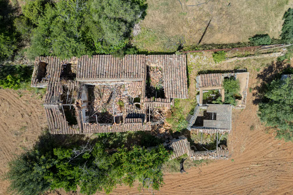 Aerial view of a dilapidated building with a red tile roof, surrounded by green trees and brown fields.