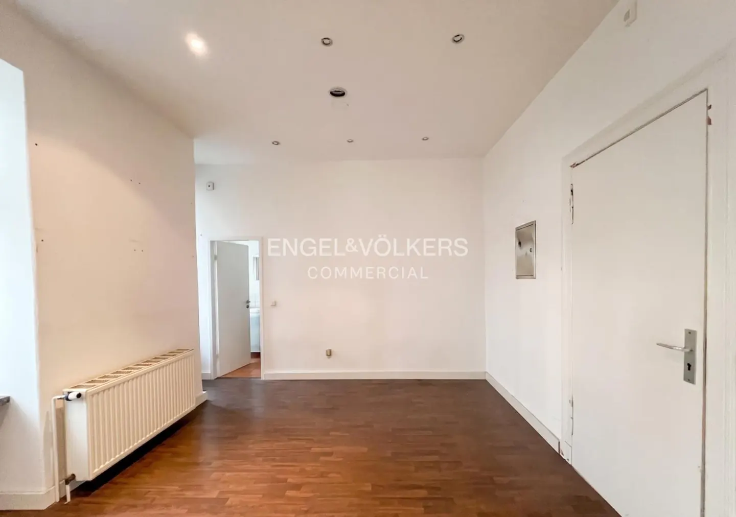 Empty room with white walls, dark wood flooring, a white radiator, and a closed door, illuminated by recessed ceiling lights.