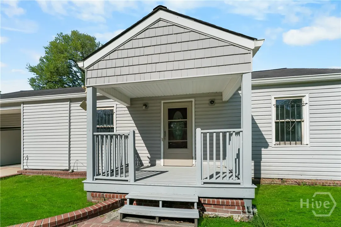 Exterior view of a light gray house with a covered porch, gray railings, and a glass-paneled front door. Green lawn in front.